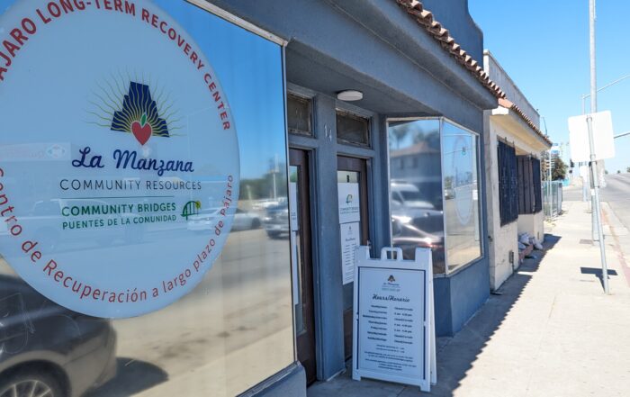 The window of the Pajaro Long-Term Recovery Center in Pajaro show the center's logo. A sandwich board sits on the sidewalk showing the center's open hours.