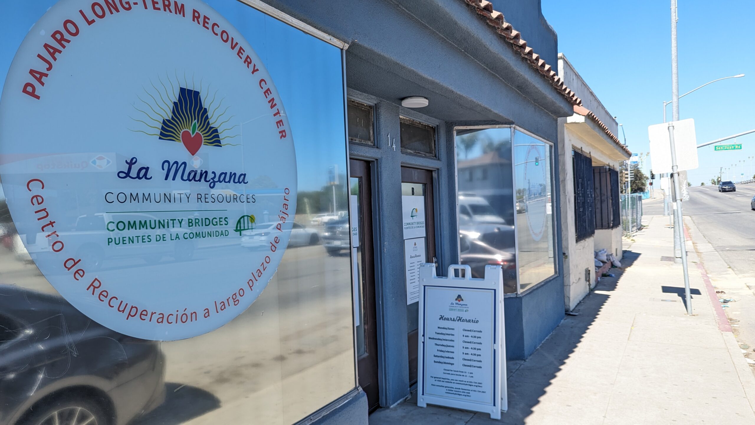 The window of the Pajaro Long-Term Recovery Center in Pajaro show the center's logo. A sandwich board sits on the sidewalk showing the center's open hours.