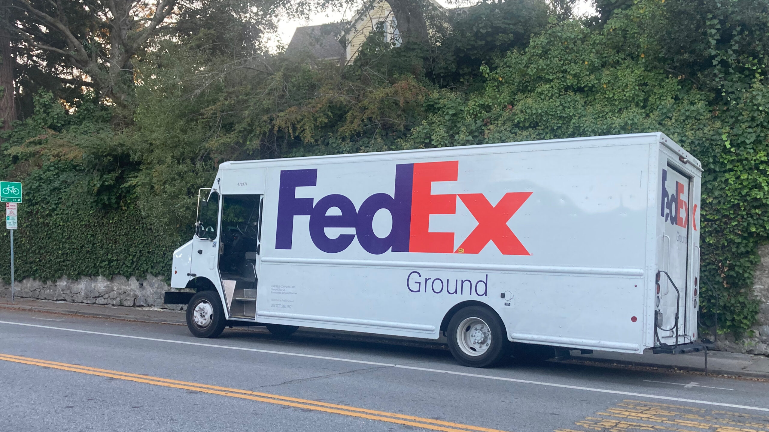 A FedEx truck is parked on an uphill street in front of a wall covered in ivy