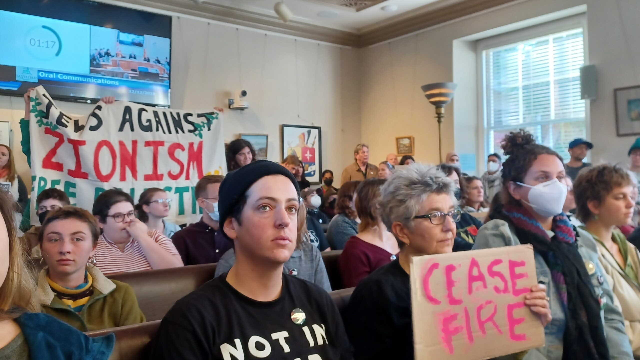 Residentes sentados en la cámara del Ayuntamiento de Santa Cruz el 12 de diciembre con pancartas en las que se leía &quot;Alto el fuego&quot; y &quot;Judíos contra el sionismo&quot;.