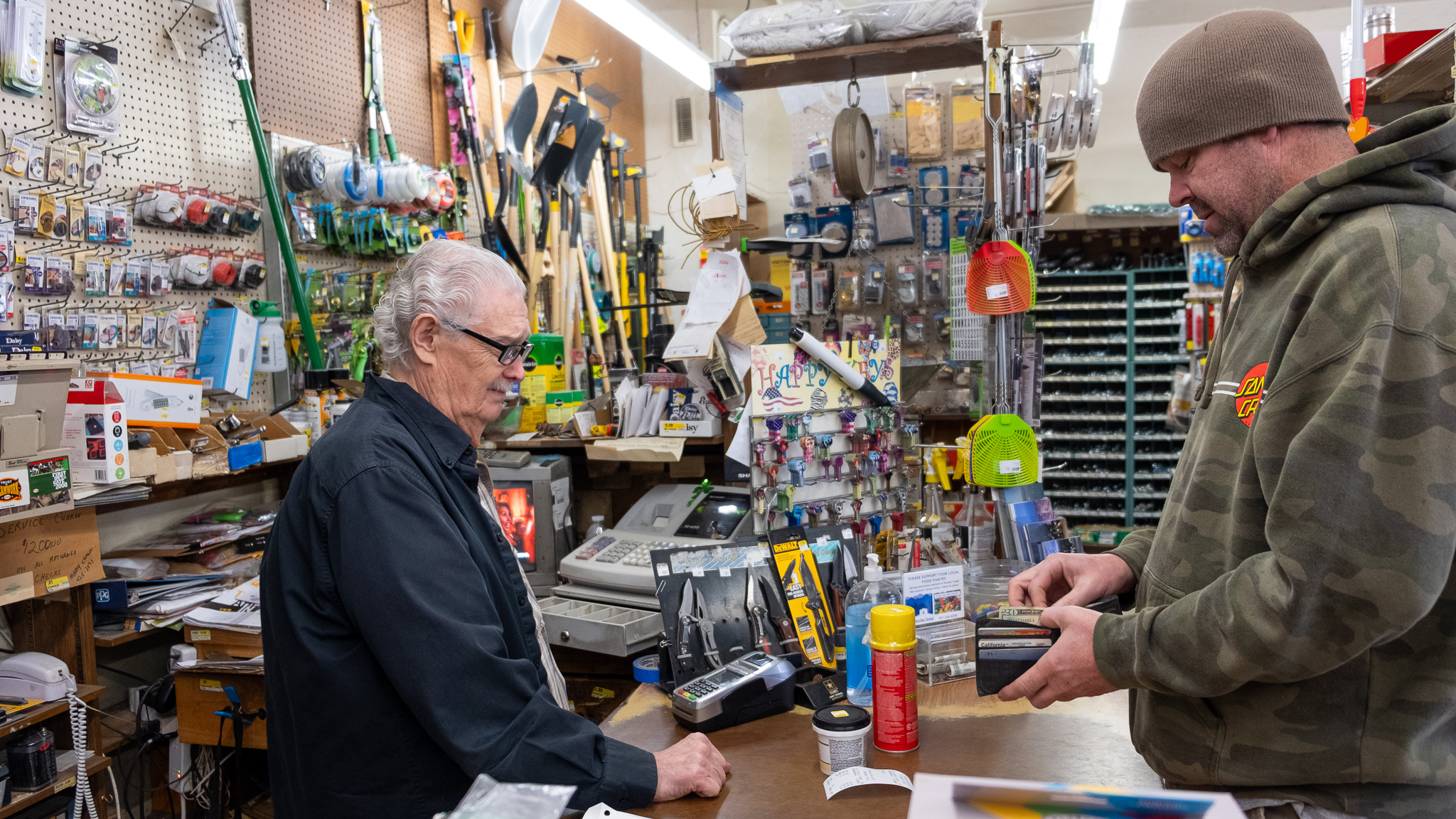 Don Walker, owner of Felton True Value Paint & Hardware on Highway 9, makes a sale. Walker has curly gray hair and black glasses, his customer is wearing a camo hoodie and a green beanie.