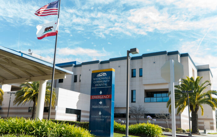 The Watsonville Community Hospital with a blue sky and a flag pole in front with the U.S. and California state flags.