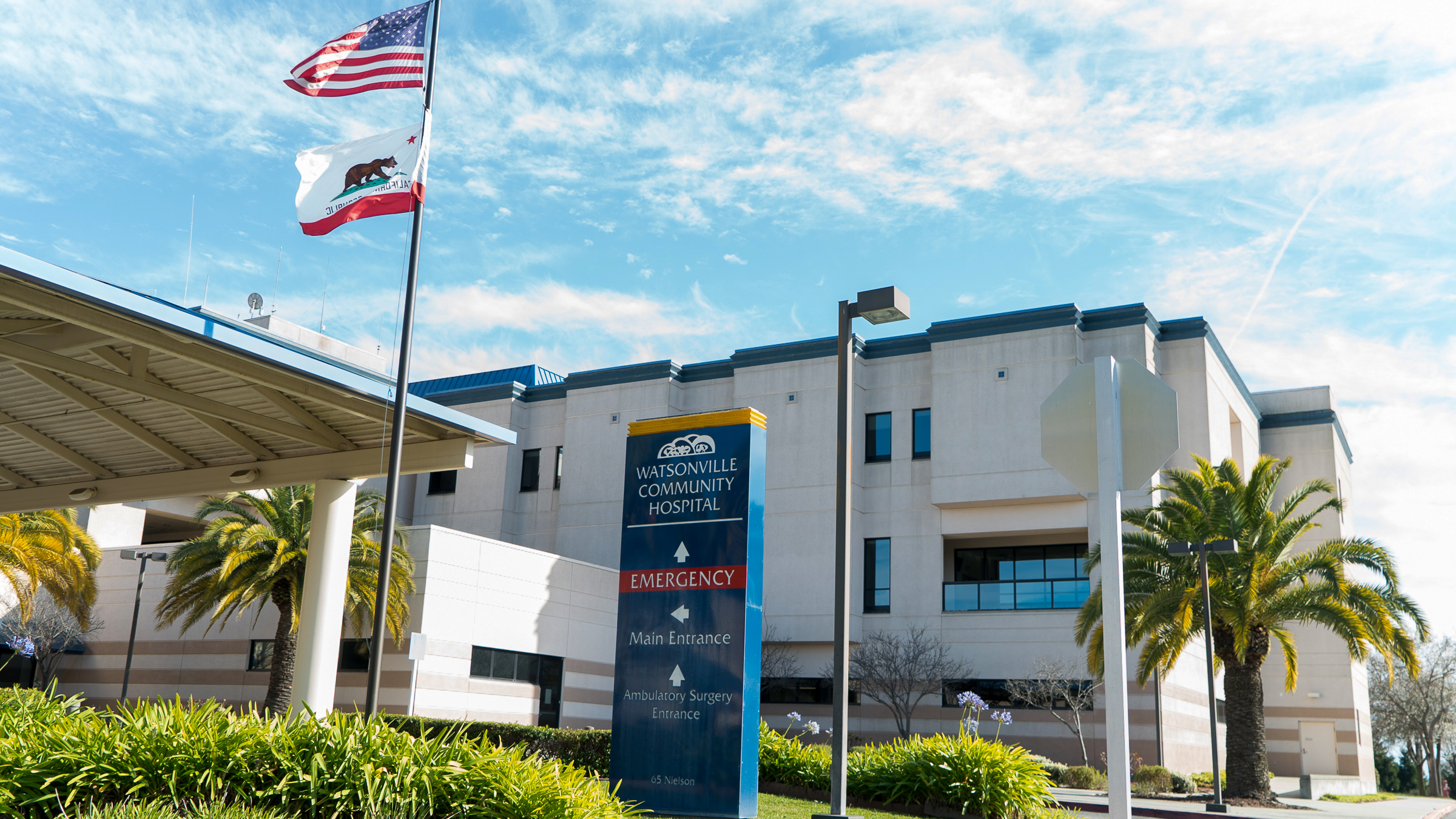 The Watsonville Community Hospital with a blue sky and a flag pole in front with the U.S. and California state flags.