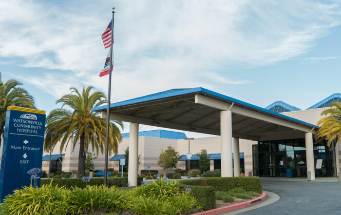 The Watsonville Community Hospital with a blue sky and a flag pole in front with the U.S. and California state flags.