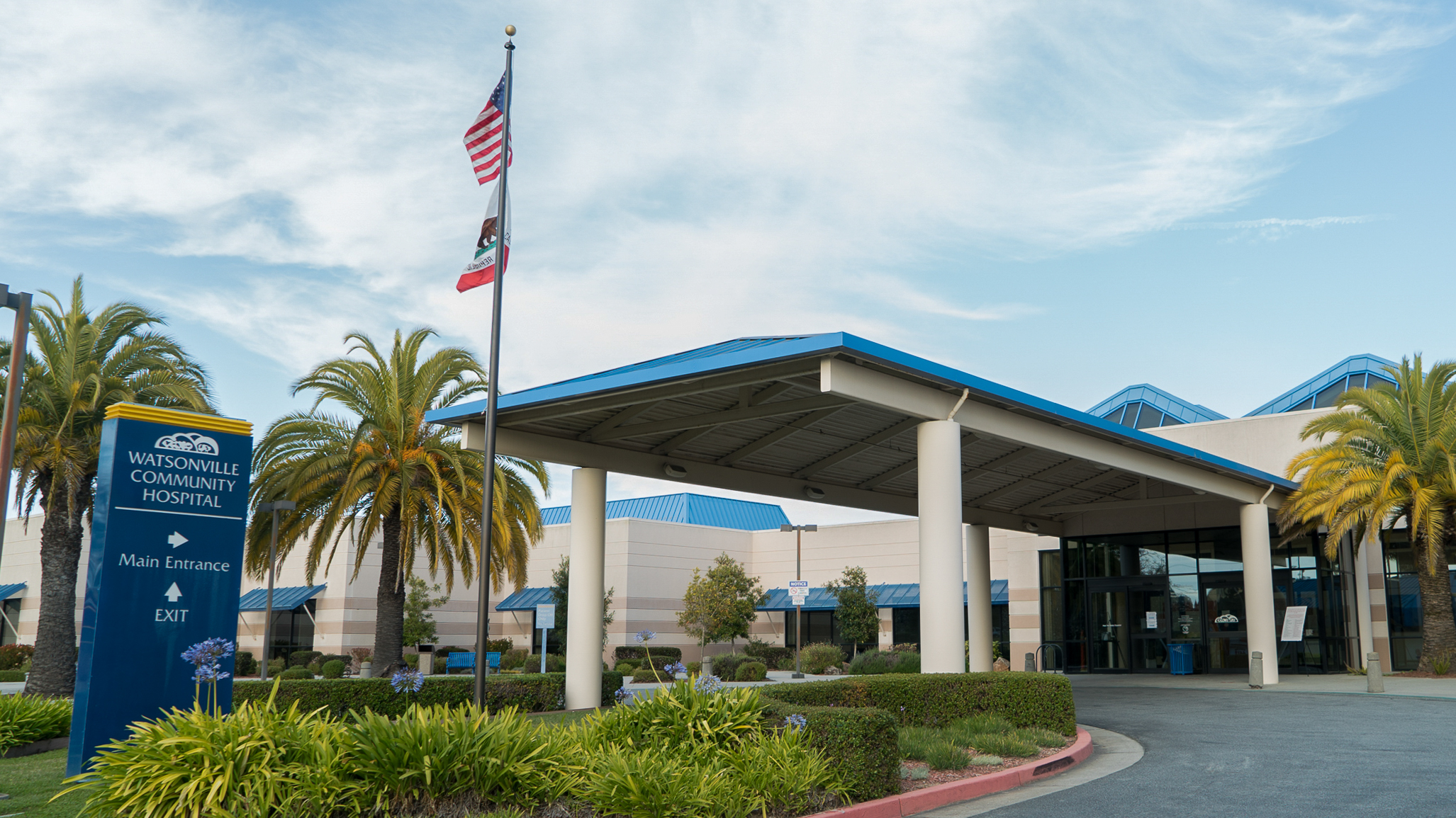 The Watsonville Community Hospital with a blue sky and a flag pole in front with the U.S. and California state flags.