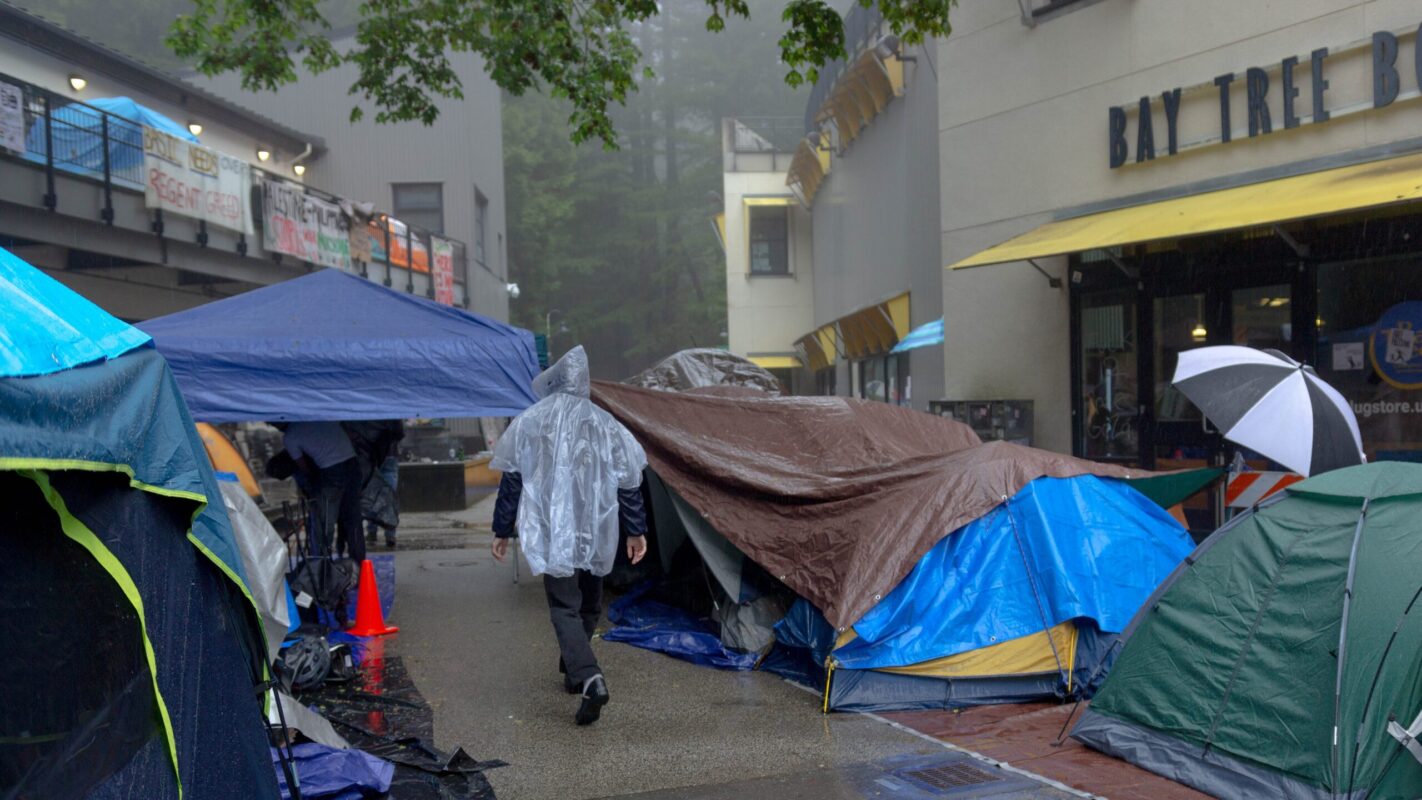Bay Tree UCSC gaza camp May 2024 Marcello Hutchinson-Trujillo 16 9 A student protestors wearing a disposable rain poncho walks between tents on a rainy day in Quarry Plaza.