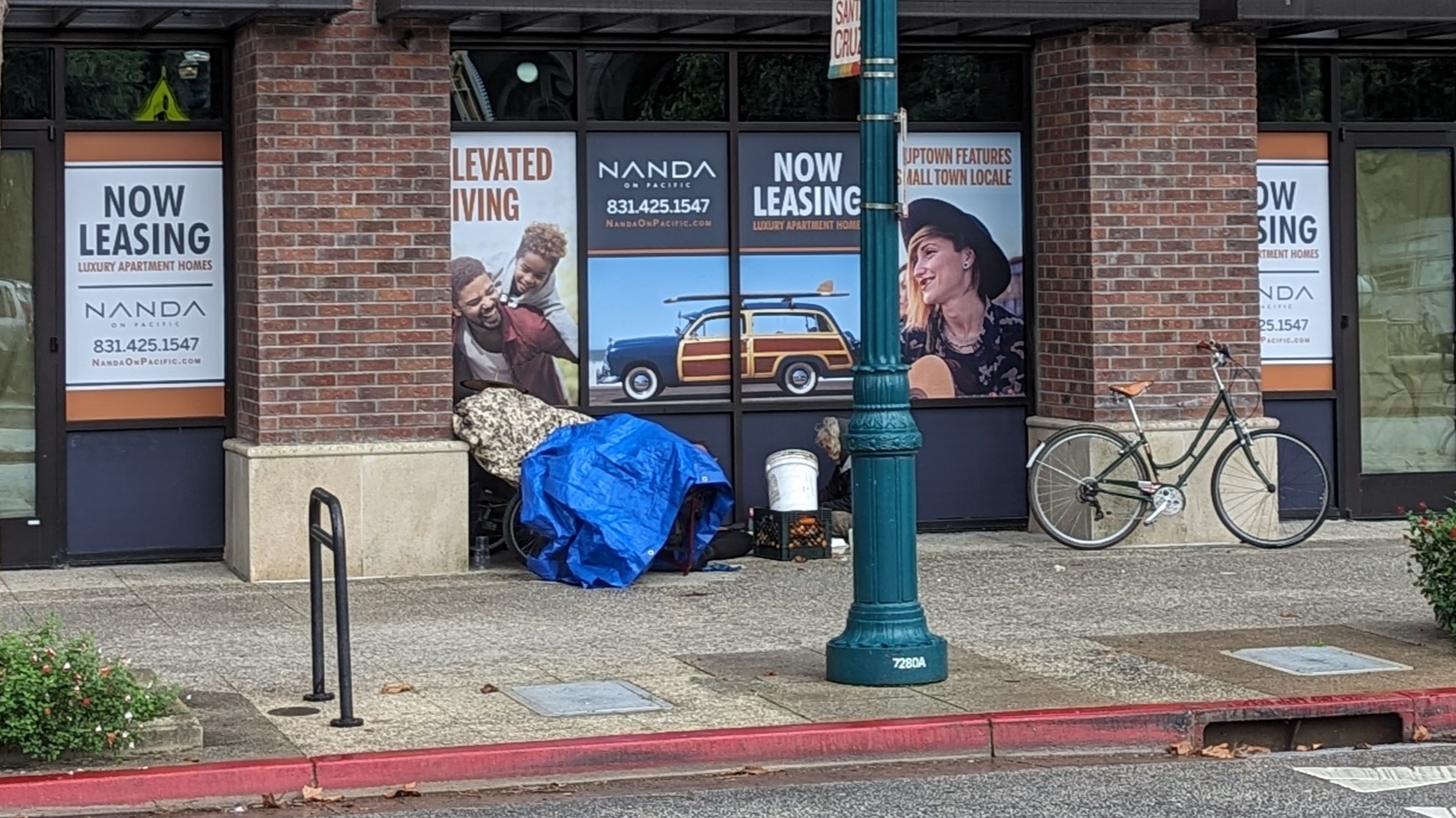 A photo showing a tarp set up on a sidewalk, under the overhang of building. The building's windows are covered in advertisements for housing that say "Now Leasing."