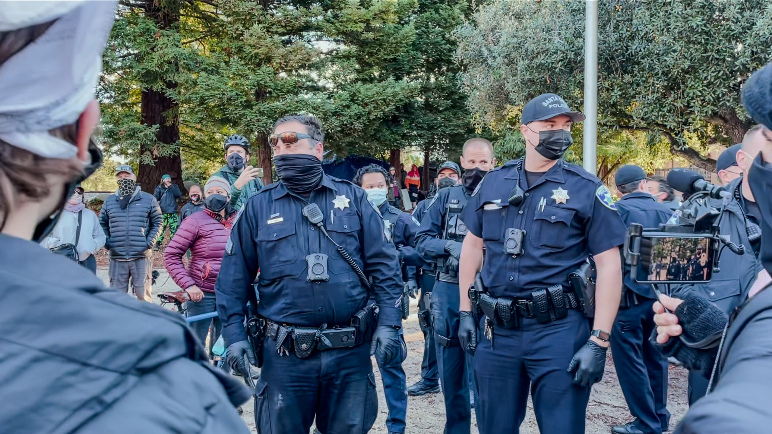 Several Santa Cruz police officers stand and speak with residents and activists during a sweep of tent camps at San Lorenzo Park in 2020.