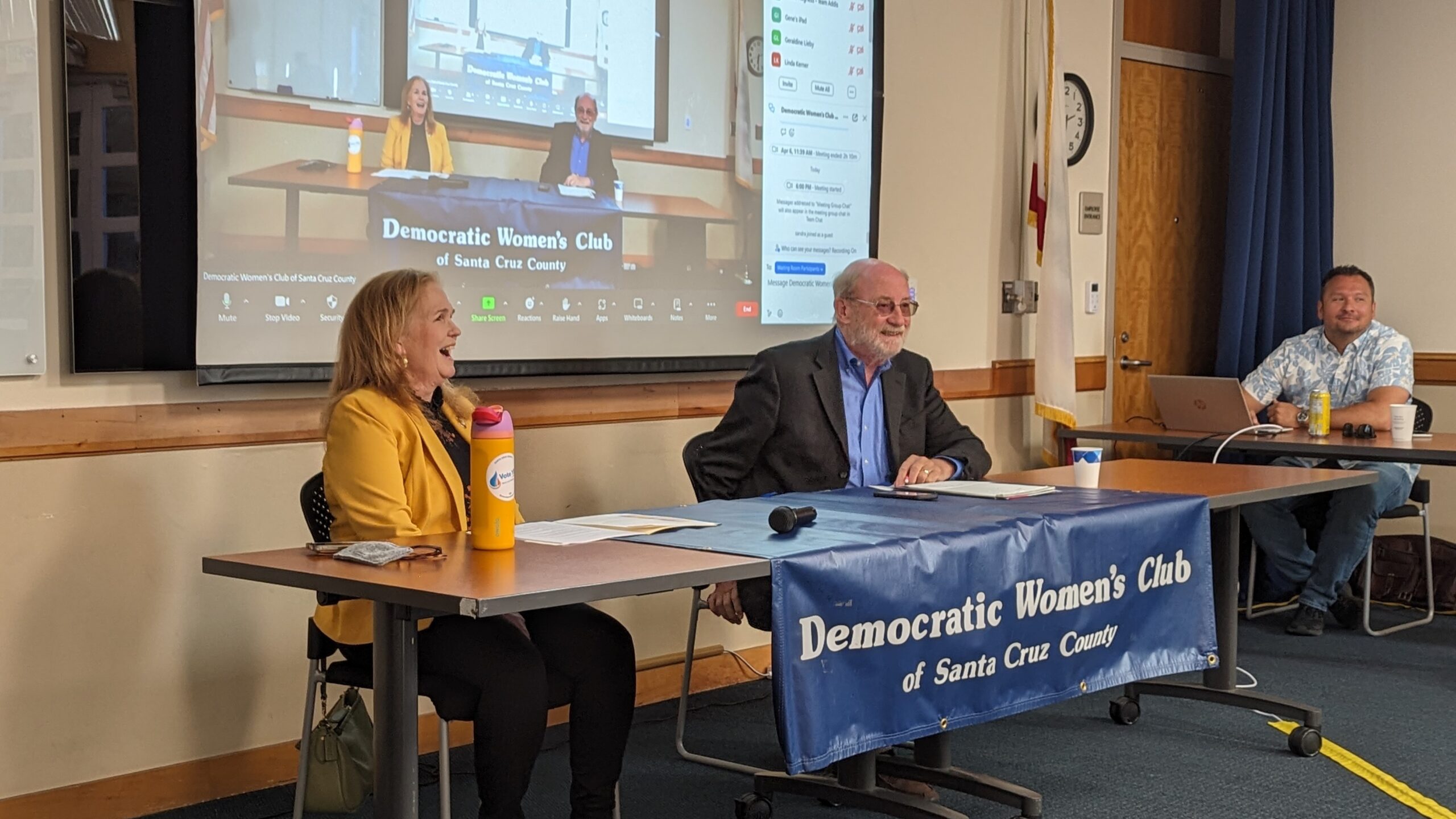 A photo of Gail Pellerin and John Laird sitting at a table with a banner hanging from the table reading "Democratic Women's Club of Santa Cruz County."