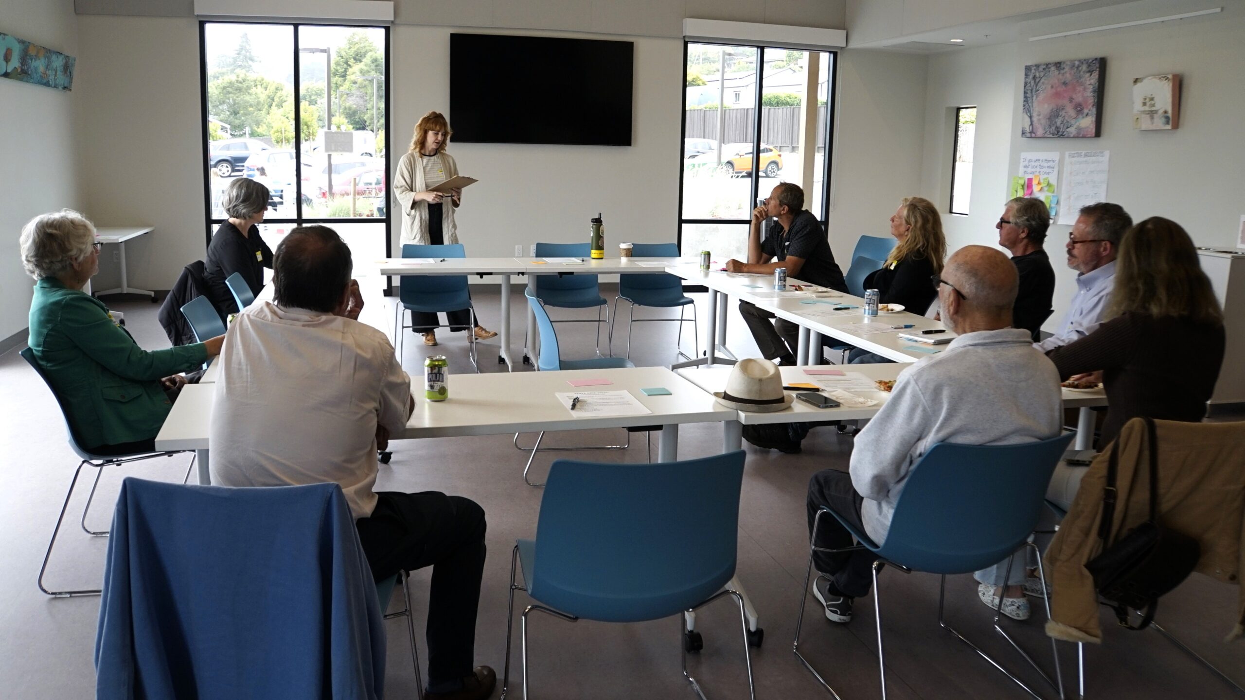 Nine people sit around tables listening as a woman stands and is speaking, holding a clip board.