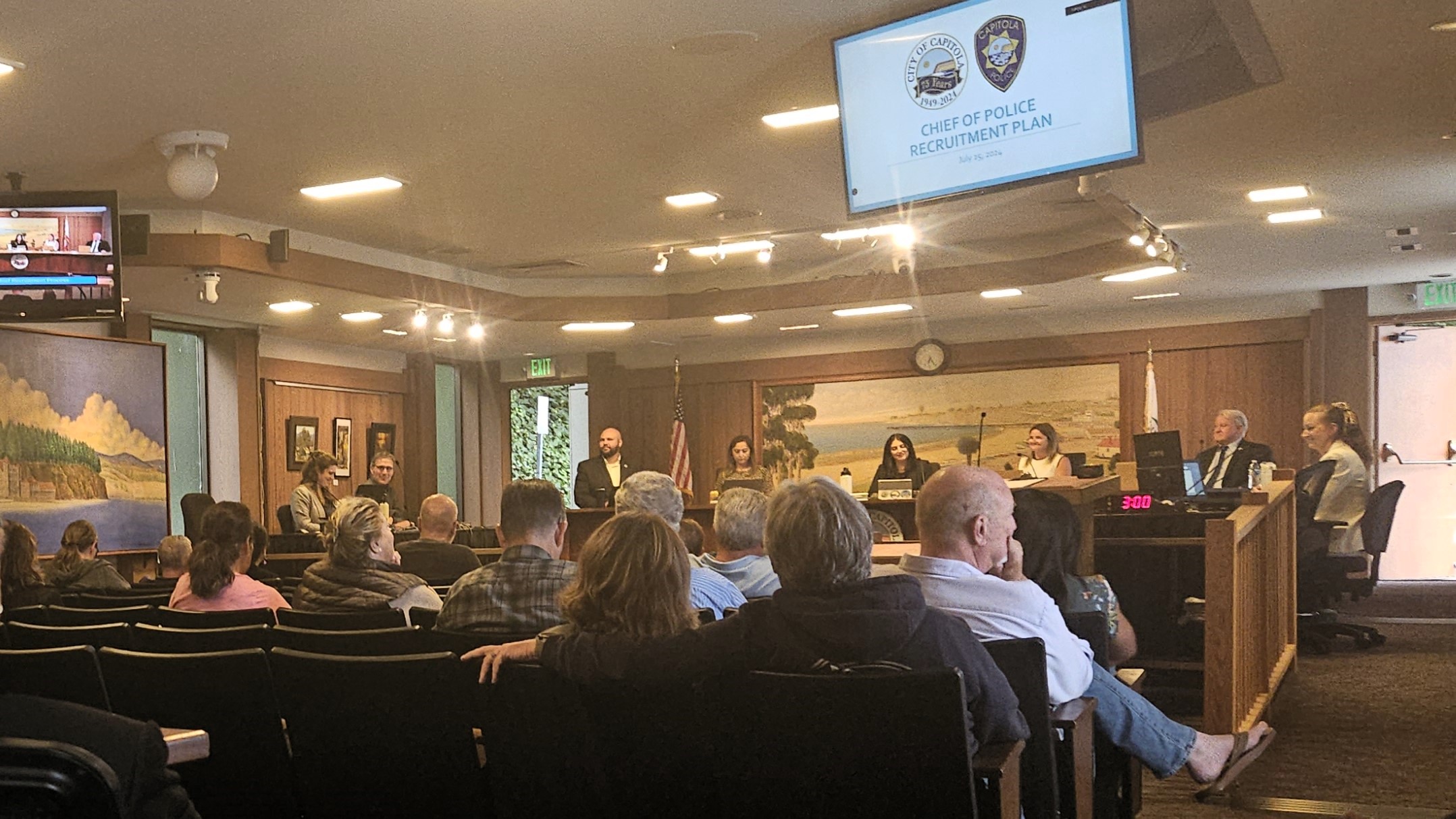 A dozen or so people sit in chairs in the Capitola City Council Chambers with council members sitting on the dais. A TV screen shows a powerpoint presentation and reads"Chief of Police Recruitment Plan."