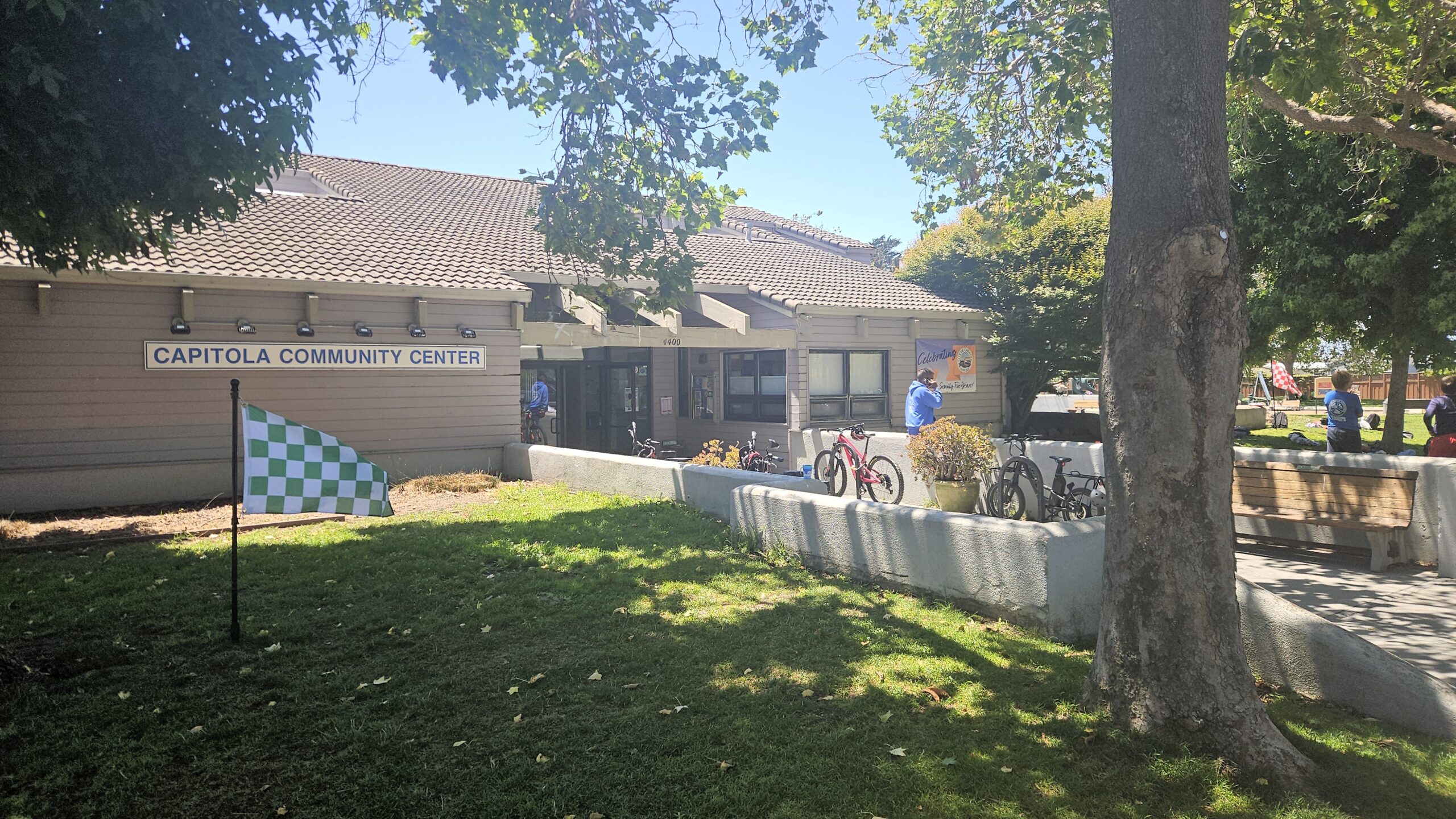 A building with a sign reading "Capitola Community Center" sits in front of a grassy area. There is a checkered green and white flag stuck in the grass.