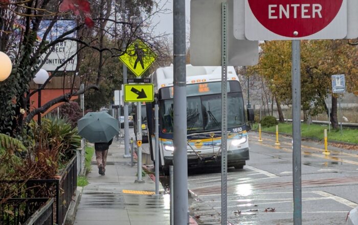 A person walks down a sidewalk with an umbrella as a Santa Cruz METRO bus pulls up on a rainy day.