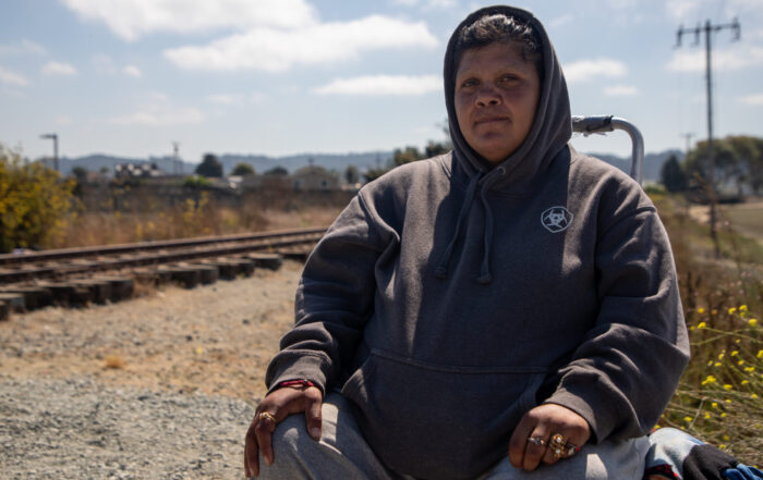 A woman wears a grey hoodie and sits on a small crate with a blue sky and clouds in the background.