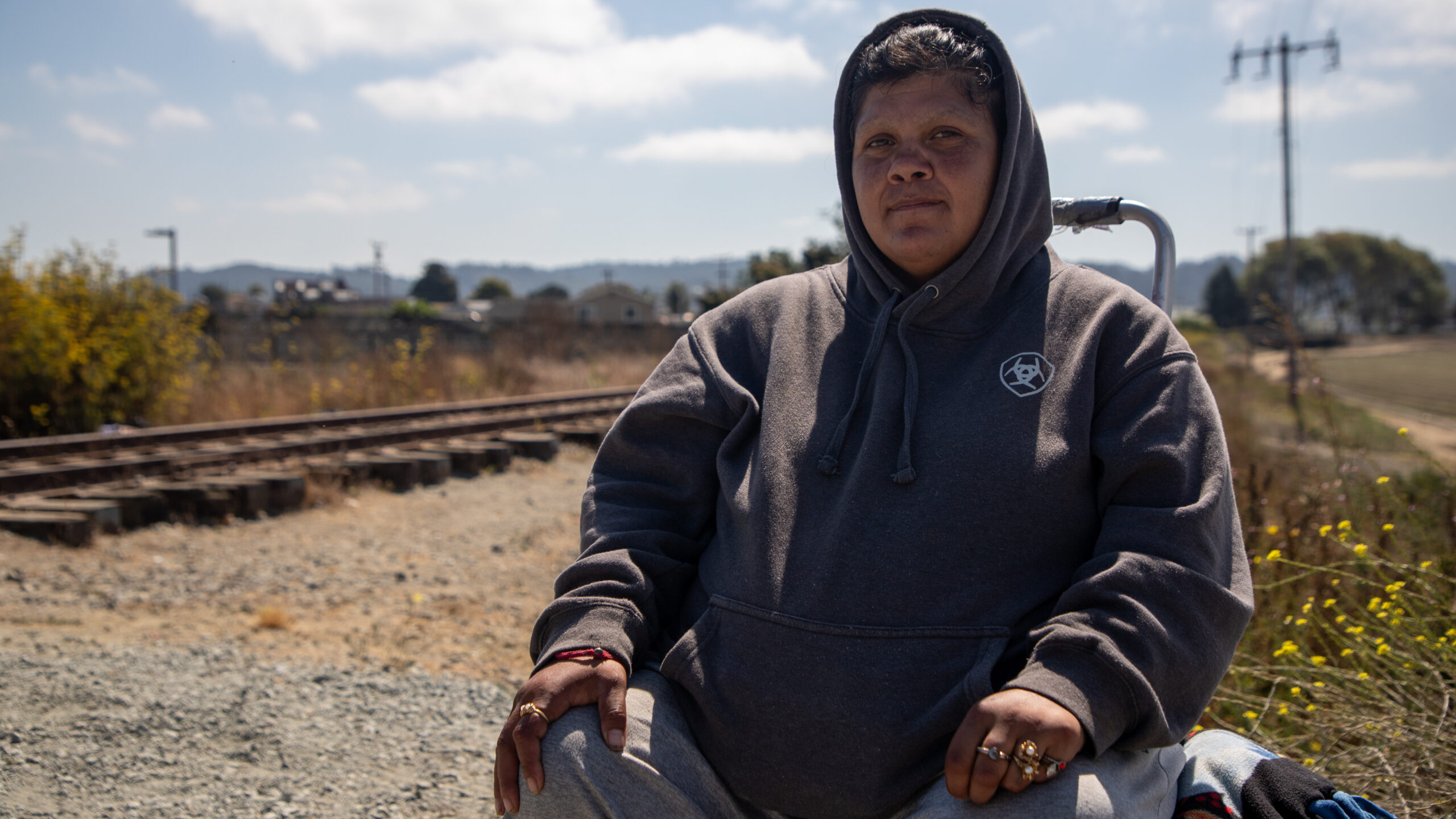 A woman wears a grey hoodie and sits on a small crate with a blue sky and clouds in the background.